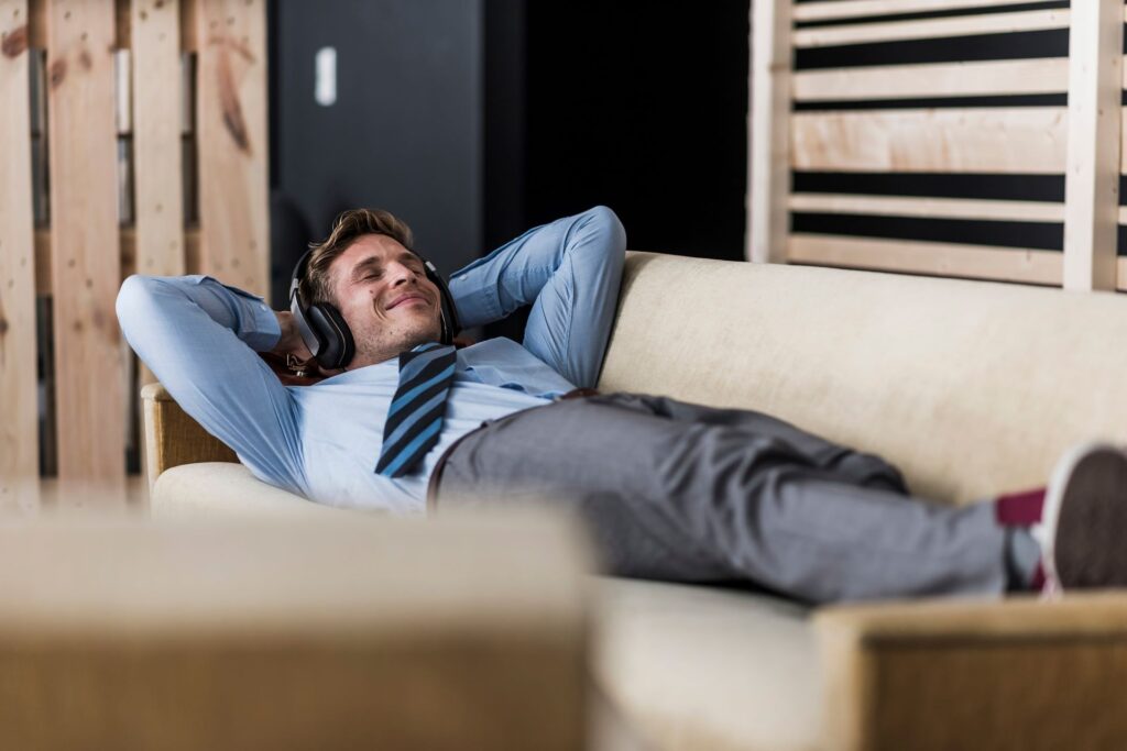 Geschäftsmann mit Kopfhörern auf der Couch in der Bürolounge liegend