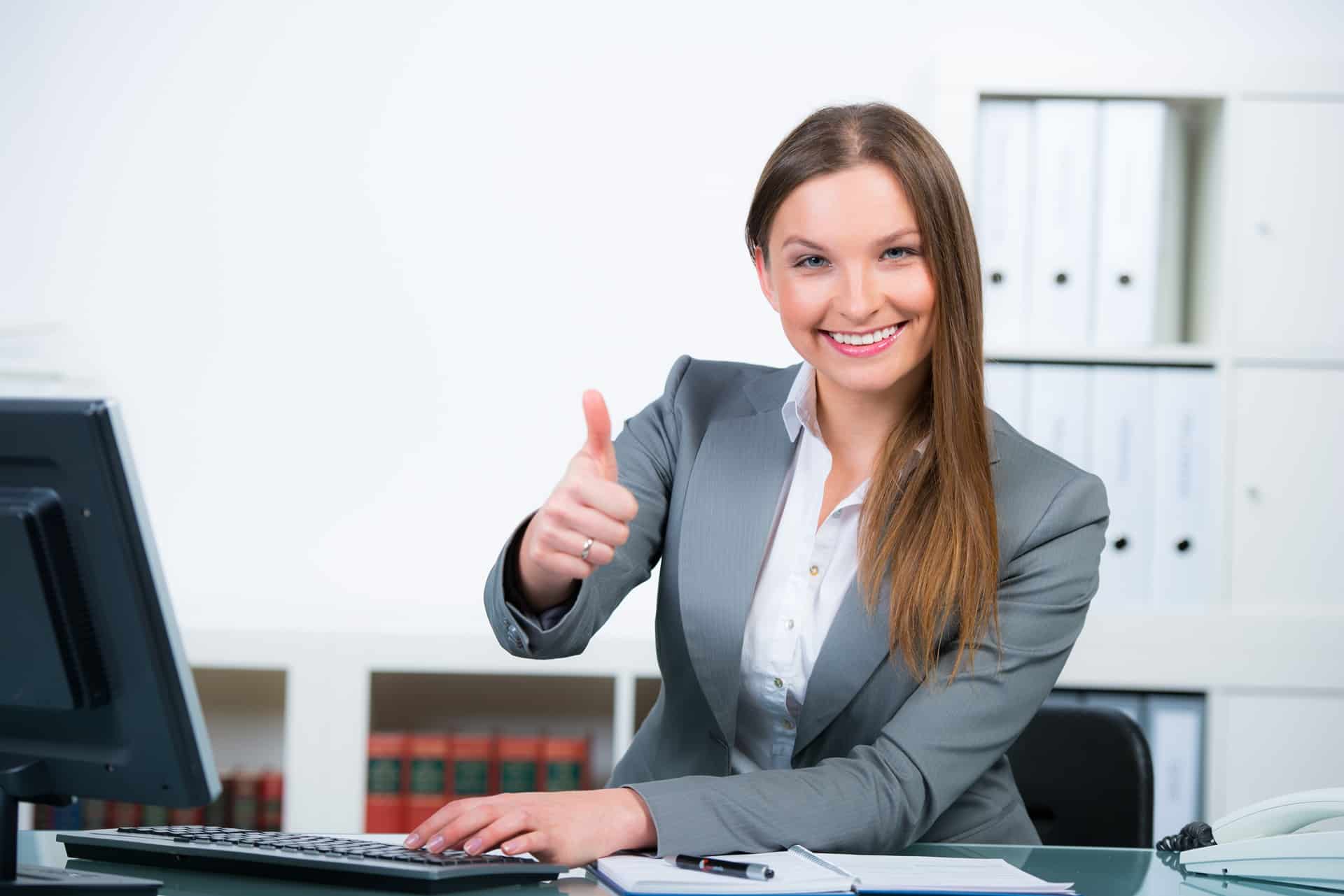 Lächelnde Frau zeigt Daumen hoch am Kanzleischreibtisch | Rechtsanwaltsfachangestellte Würzburg